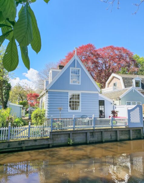 A serene blue house by the tranquil waters of a river, reflecting the sky and surrounded by lush greenery A serene blue house by the tranquil waters of a river, reflecting the sky and surrounded by lush greenery. Broek in Waterland Netherlands