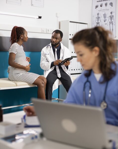 Doctor consults with pregnant patient Physician in lab coat gives prenatal advice to pregnant lady while nurse sits at nearby desk reviewing medical files. Female healthcare worker reads patient records while doctor consults with patient