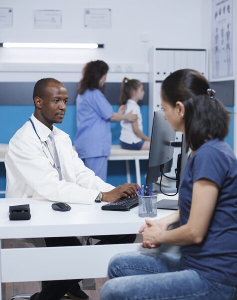 Doctor discussing with female patient In medical office, African American doctor discusses with caucasian lady about healthcare and medicine. Black man using the desktop PC, talks to the patient while wearing a lab coat and mask.