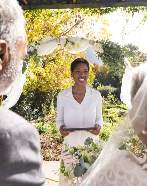 Happy biracial female marriage officiant with tablet and senior couple at wedding in sunny garden. Love, ceremony, marriage, summer, communication and celebration, unaltered. Happy biracial female marriage officiant with tablet and senior couple at wedding in sunny garden. Love, ceremony, marriage, summer, communication and celebration, unaltered.