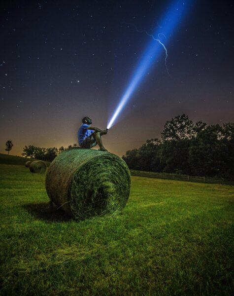 pexels-photo-1816699-1816699 A man sits on a hay bale in a field, shining a flashlight into the night sky full of stars.